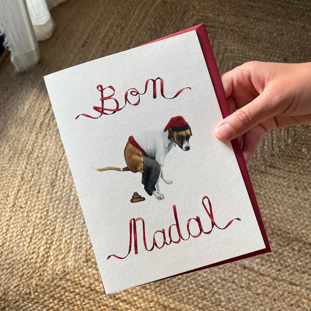Hand holding a christmas card that says "Bon Nadal" with a handpainted waterolor illustration of a dog pooping while wearing the traditional catalan farmers clothes, in the caganer style