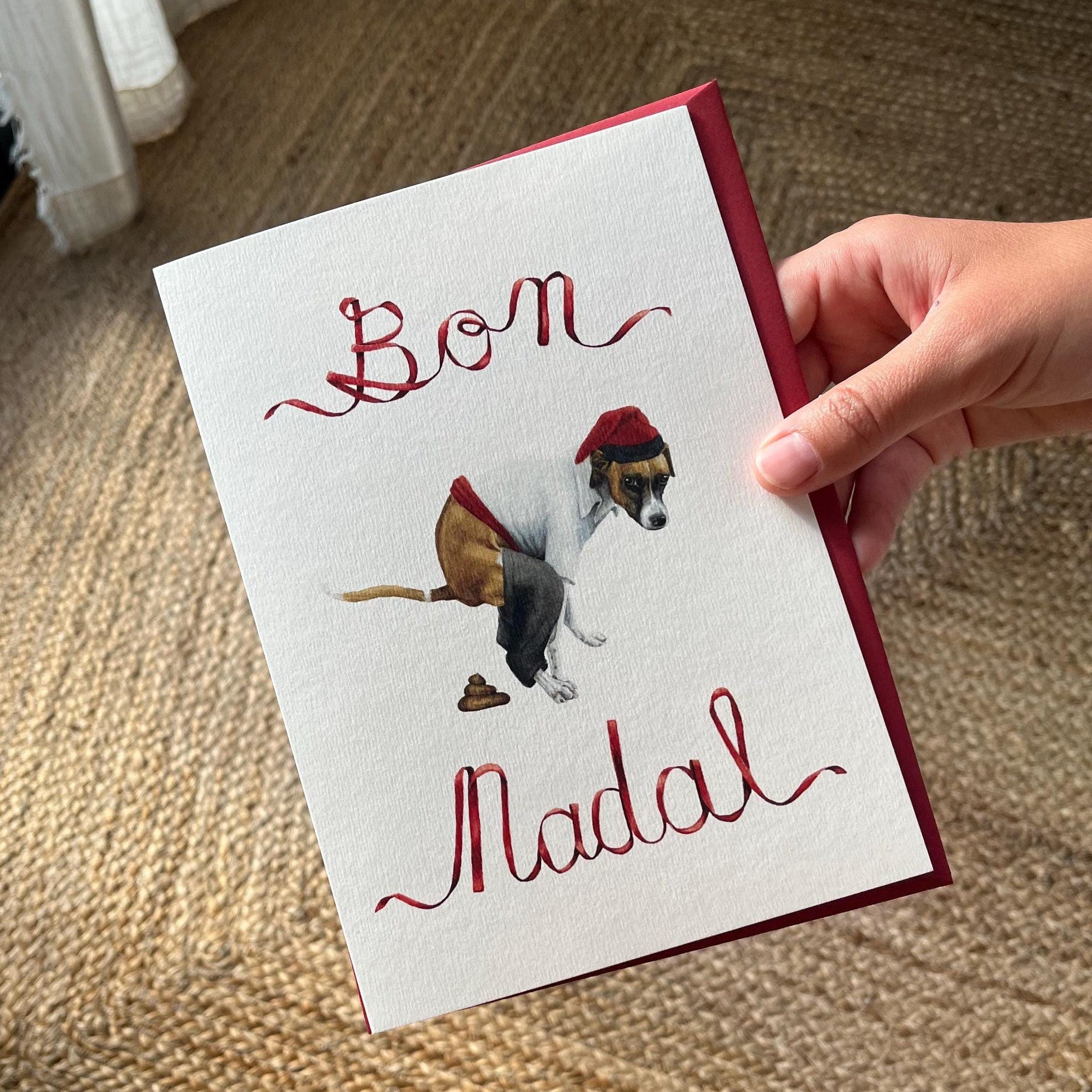 Hand holding a christmas card that says "Bon Nadal" with a handpainted waterolor illustration of a dog pooping while wearing the traditional catalan farmers clothes, in the caganer style
