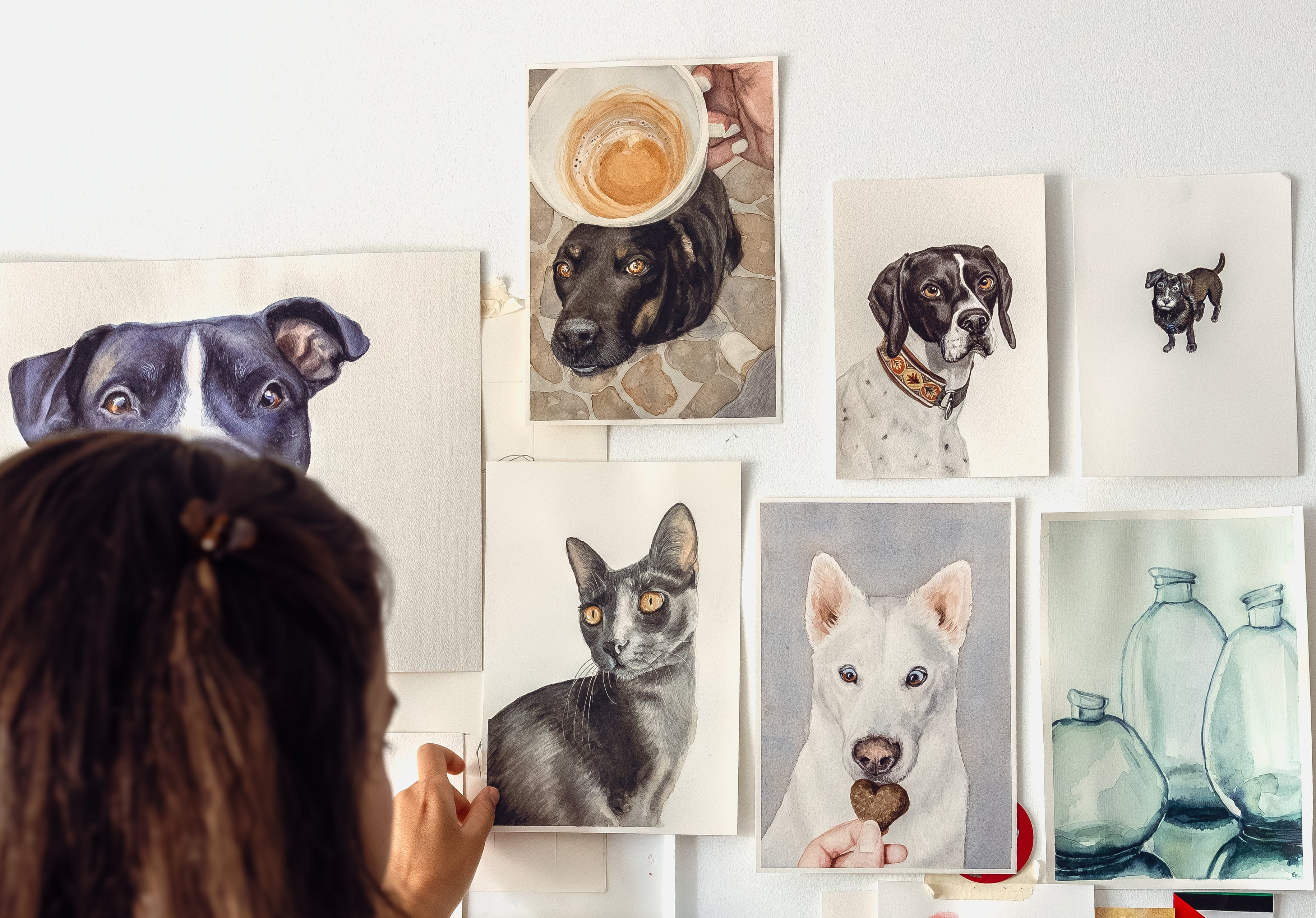 the back of a woman's head as she adjusts a painting on a white wall full of handpainted watercolor pet portraits. There are 5 dog paintings, 1 cat painting and a painting of three glass bottles.